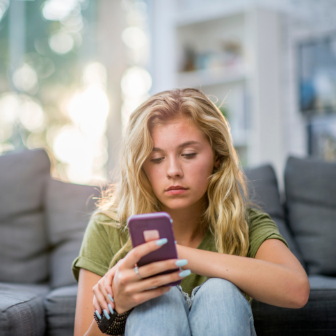 Eine junge Frau sitzt auf dem Boden und lehnt sich an ein Sofa an. SIe shcaut auf ihr Smartphone.