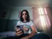 Das Foto zeigt eine junge Person, die in einem abgedunkelten Raum sitzt. Sie hat ein Smartphone in der Hand, auf das sie schaut. Ihr Blick ist ernst. Das Foto ist von schräg unten aufgenommen, sodass das Smartphone im Vordergrund zu sehen ist und der Oberkörper und das Gesicht der Person dahinter.