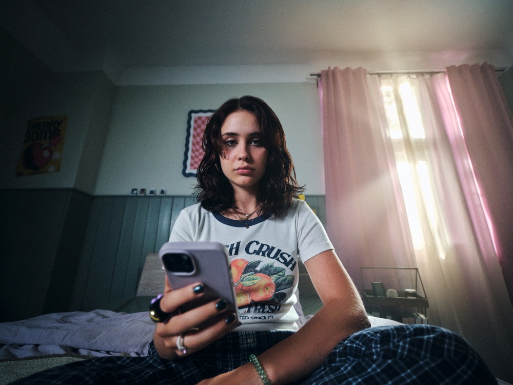 Das Foto zeigt eine junge Person, die in einem abgedunkelten Raum sitzt. Sie hat ein Smartphone in der Hand, auf das sie schaut. Ihr Blick ist ernst. Das Foto ist von schräg unten aufgenommen, sodass das Smartphone im Vordergrund zu sehen ist und der Oberkörper und das Gesicht der Person dahinter.